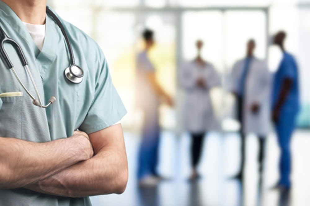 A close-up of a healthcare professional in scrubs with arms crossed and a stethoscope around their neck, with a blurred background of medical staff in a hospital or clinic setting.