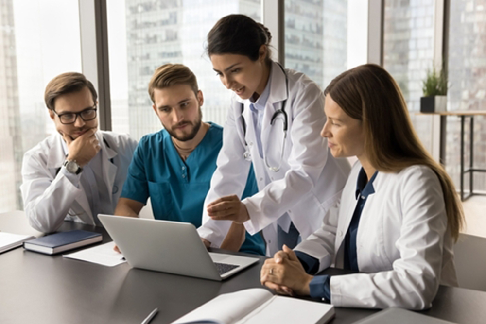 Young Indian medical team leader presenting job application on laptop to colleagues. Diverse doctors meeting at computer, talking on group video call, looking at screen, speaking, listening