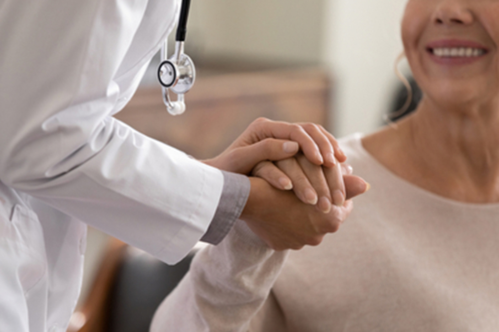 Doctor giving hope. Close up shot of young female physician leaning forward to smiling elderly lady patient holding her hand in palms. Woman caretaker in white coat supporting encouraging old person